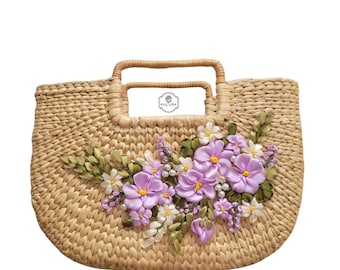 May include: A straw tote bag with a woven design and a floral embellishment. The flowers are made of purple and white ribbon and are accented with pearl beads. The bag has two woven handles.