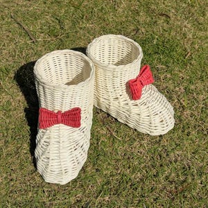 May include: Two woven wicker boot-shaped planters. The planters are off-white with a red bow on the front of each boot. The planters are sitting on green grass.