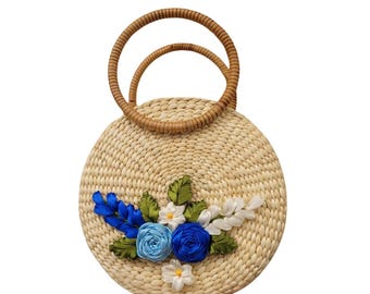 May include: Round woven straw handbag with two brown handles. The bag is decorated with blue, white, and green fabric flowers. The flowers are arranged on the front of the bag.