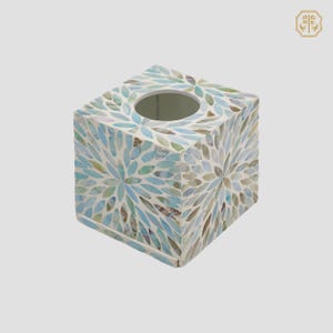 Puede incluir: Caja de pañuelos cuadrada con diseño de mosaico. La caja está cubierta de pequeñas baldosas iridiscentes en tonos azules, verdes y blancos, dispuestas en un patrón floral. Tiene una abertura circular para dispensar pañuelos. Un artículo decorativo.