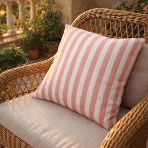 May include: A square decorative pillow with vertical stripes in light pink and white rests on a wicker chair. The chair has a beige cushion. The setting is outdoors, with plants and a stone wall in the background.