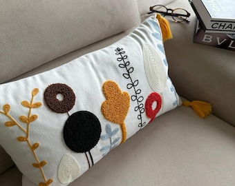 May include: A white throw pillow with embroidered colorful shapes and tassels. The pillow features a black circle, a brown circle, a yellow heart, a red circle, and blue and yellow leaves.