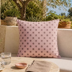 May include: A square throw cushion featuring a light pink fabric base with a repeating pattern of evenly spaced, solid dark grey polka dots. The cushion has a clean, finished edge and a soft, matte texture, presenting a simple and uniform design.