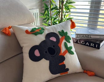 May include: A white throw pillow with a black and orange embroidered koala bear design. The koala is holding onto a branch with green leaves. The pillow has orange tassels on the corners.
