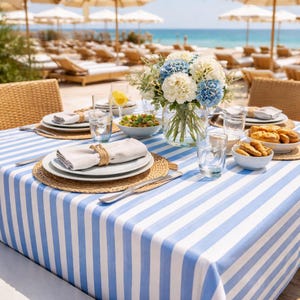 May include: A table set for a beachside meal, featuring a blue and white striped tablecloth. The table is set with plates, napkins, glasses, and a bouquet of white and blue flowers. The background shows a beach with lounge chairs and umbrellas.