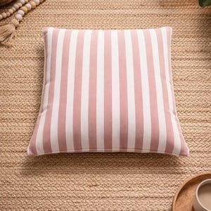 May include: Square decorative pillow with vertical stripes in white and light pink. The pillow is centered on a woven jute rug. The pillow is a home decor item.