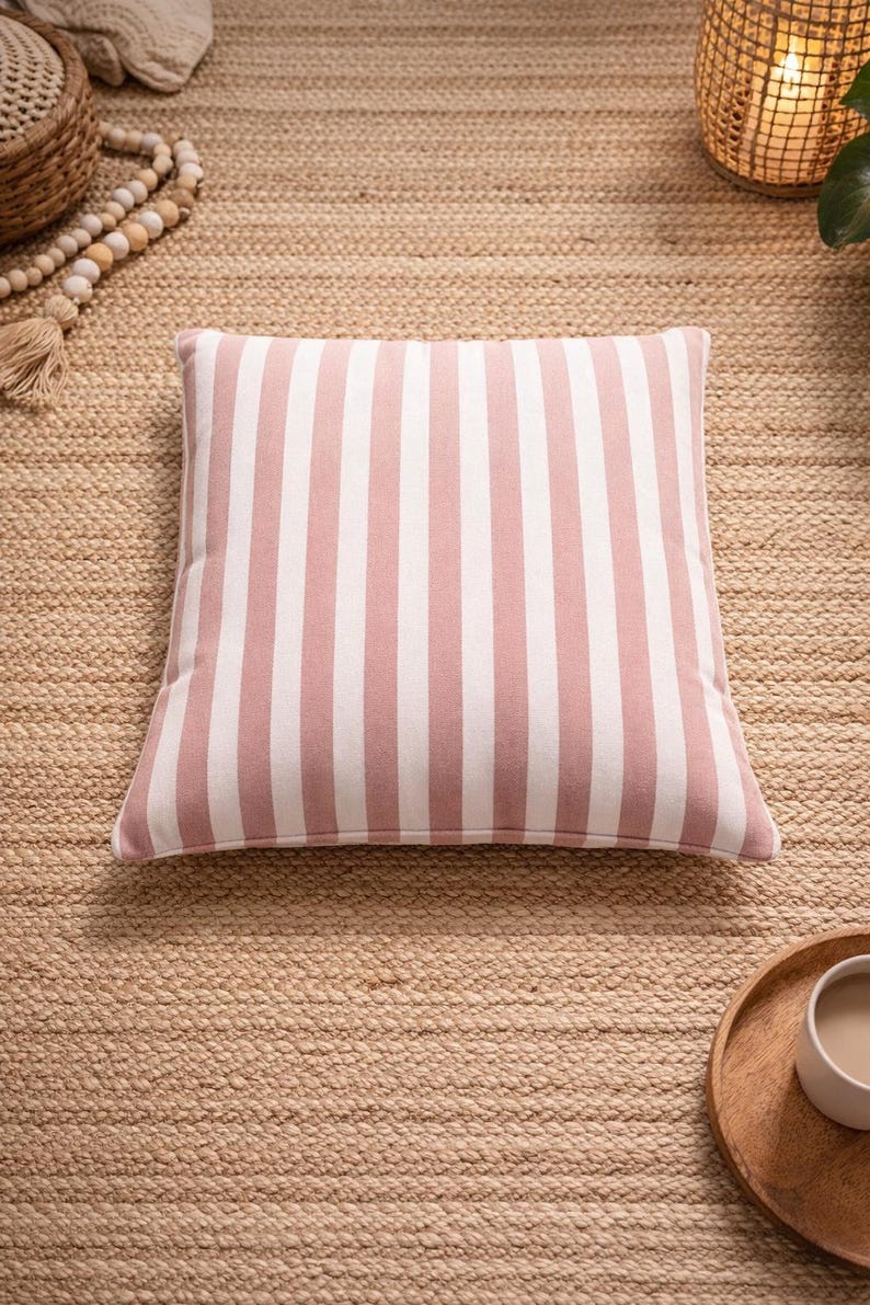 May include: Square decorative pillow with vertical stripes in white and light pink. The pillow is centered on a woven jute rug. The pillow is a home decor item.