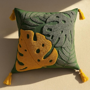 Pode incluir: Almofada quadrada verde com folhas de monstera bordadas em amarelo e verde. A almofada tem borlas amarelas em cada canto. O design apresenta uma grande folha amarela e folhas verdes menores.