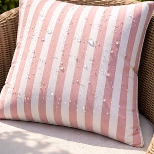 May include: A square outdoor pillow with a pink and white striped pattern. Water droplets are visible on the fabric. The pillow is placed on a wicker chair with a white cushion. The pillow is designed for outdoor use.