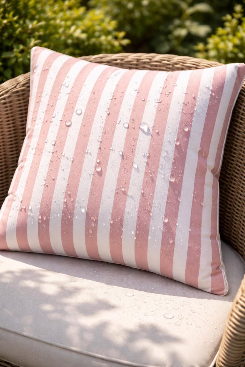 May include: A square outdoor pillow with a pink and white striped pattern. Water droplets are visible on the fabric. The pillow is placed on a wicker chair with a white cushion. The pillow is designed for outdoor use.