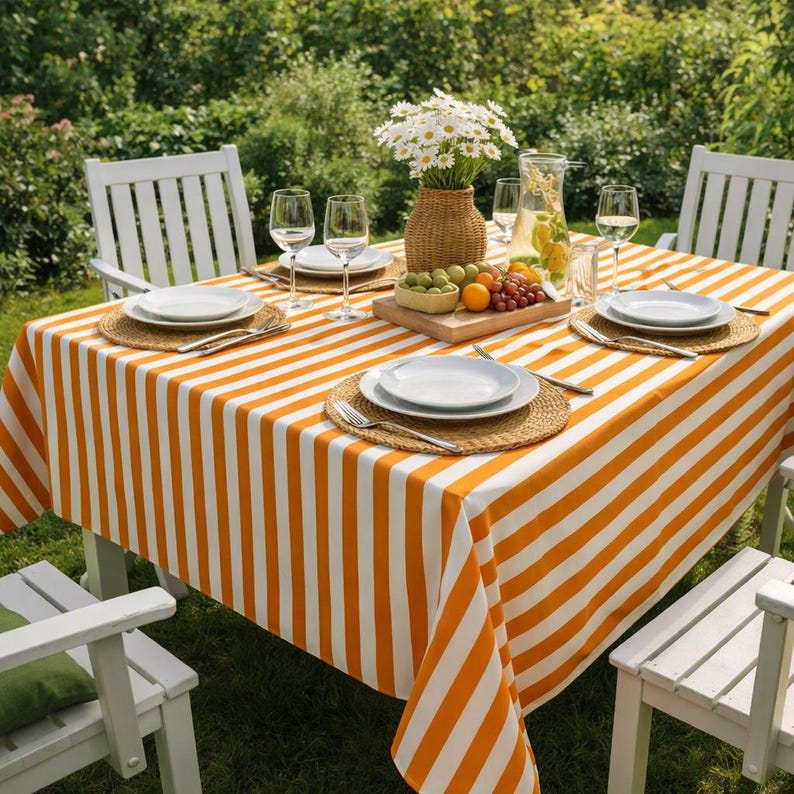 May include: An outdoor dining table with an orange and white striped tablecloth. The table is set with white plates, woven placemats, wine glasses, and a vase of white flowers. Fresh fruit and drinks are also present.