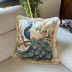 Könnte beinhalten: Dekoratives quadratisches Kissen mit detailliertem Pfauendesign. Das Kissen hat einen cremefarbenen Hintergrund mit einem blauen, türkisfarbenen und goldenen Pfau. Das Kissen hat einen Fransenrand und liegt auf einem beigen Sofa.
