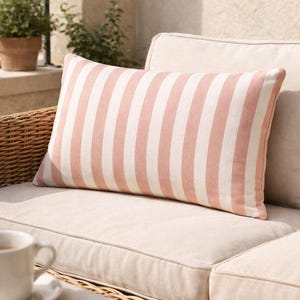 May include: A rectangular pillow with vertical stripes in light pink and white. The pillow is placed on a beige outdoor sofa with a woven wicker frame. A cup of coffee sits on a nearby table.
