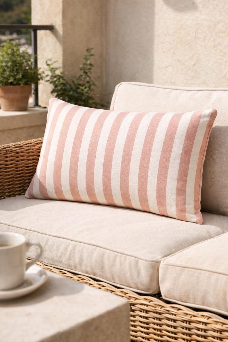 May include: A rectangular pillow with vertical stripes in light pink and white. The pillow is placed on a beige outdoor sofa with a woven wicker frame. A cup of coffee sits on a nearby table.