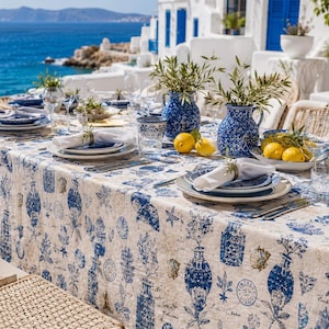 May include: A rectangular dining table covered in a beige tablecloth featuring a blue botanical and vase print. The table is set with white plates, blue patterned bowls, clear glassware, white napkins, and two blue ceramic pitchers with greenery.