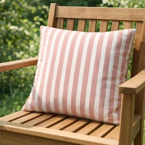 May include: A square outdoor pillow with vertical stripes in white and a muted rose color rests on a wooden chair. The chair is made of light brown wood and is positioned outdoors in a garden setting.