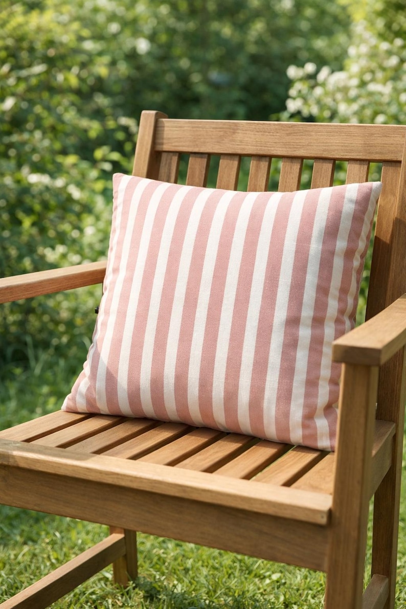 May include: A square outdoor pillow with vertical stripes in white and a muted rose color rests on a wooden chair. The chair is made of light brown wood and is positioned outdoors in a garden setting.