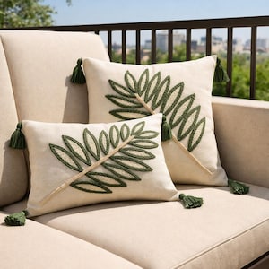 Puede incluir: Dos cojines decorativos color crema con un diseño de hojas verdes bordadas. Las hojas están bordadas con una puntada texturizada. Cada cojín tiene borlas verdes en las esquinas. Los cojines están sobre un sofá beige.