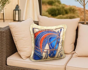 May include: A decorative square pillow featuring a blue and red horse head design. The pillow has a cream-colored fringe border and is placed on a beige sofa with other pillows. The horse design is detailed with blue and red accents.