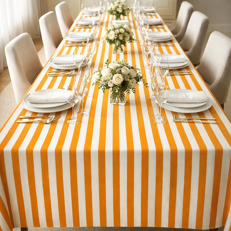 May include: A dining table set for a formal occasion. The table is covered with an orange and white striped tablecloth. White plates, napkins, and cutlery are set for each guest. Clear wine glasses and small floral arrangements complete the setting.