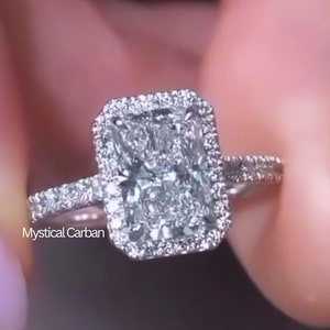 Op de afbeelding: Een zilveren ring met een grote, rechthoekige, heldere diamant omgeven door een halo van kleinere diamanten. De ring is gezet op een witte band.