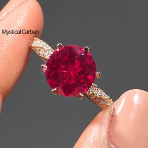 Pode incluir: Um anel de ouro rosa com uma grande pedra preciosa redonda e vermelha escura. A banda é adornada com pequenas pedras claras. O anel é segurado pelos dedos de uma pessoa. O texto "Mystical Carban" é visível.