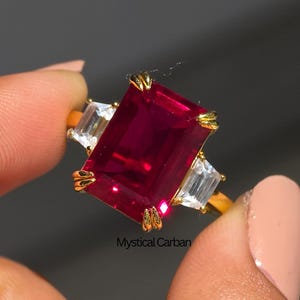 May include: A gold ring with a large, rectangular, deep red gemstone and two smaller, white, rectangular gemstones. The ring is held up against a grey background. The ring is a piece of jewellery.
