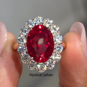 Può includere: Un anello d'oro con una grande gemma rossa intensa a taglio ovale, circondata da un alone di gemme più piccole e chiare. L'anello è tenuto su uno sfondo morbido e sfocato, evidenziando il colore vibrante e la brillantezza della pietra centrale.