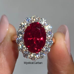 May include: A gold ring featuring a large, oval-cut, deep red gemstone surrounded by a halo of smaller, sparkling white stones. The ring is held up by a hand, with the text "Mystical Carban" visible.