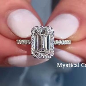 Peut inclure: Une bague en diamant avec une pierre centrale de taille émeraude et un halo de petits diamants. La monture est également ornée de petits diamants. La bague est tenue par une personne avec du vernis à ongles blanc. Le texte "Mystical Carban" est visible.