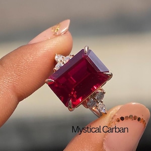 Pode incluir: Um anel de prata com uma grande pedra preciosa retangular, vermelho escuro, como ponto focal. Pedras preciosas menores e claras são colocadas ao redor da pedra principal. O anel é segurado contra um fundo desfocado.