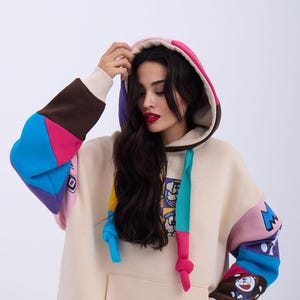 May include: A cream coloured hoodie with a colourful patchwork design. The hoodie has a large pocket on the front and a drawstring hood. The sleeves are made of different coloured fabrics, including blue, pink, purple, and brown. The hoodie features a cartoon character graphic on the front.