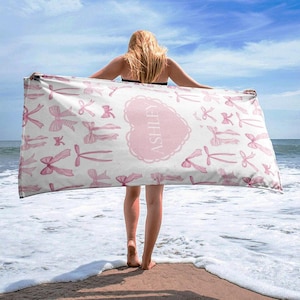 Puede incluir: Una toalla de playa blanca con lazos rosas y el nombre "Ashley" en un coraz&oacute;n rosa. La toalla est&aacute; doblada y sostenida por una persona en una playa.