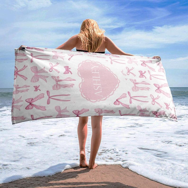 Puede incluir: Una toalla de playa blanca con lazos rosas y el nombre "Ashley" en un coraz&oacute;n rosa. La toalla est&aacute; doblada y sostenida por una persona en una playa.