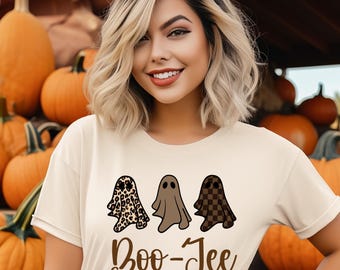 May include: A beige t-shirt featuring three cartoon ghosts with different patterns: leopard print, solid brown, and checkered. The text "Boo-Jee" is printed below the ghosts in a brown script font. Pumpkins are in the background.
