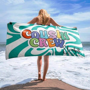 Może przedstawiać: Ręcznik plażowy z napisem "COUSIN CREW" w kolorowych literach bąbelkowych, z imieniem "Elizabeth" kursywą. Ręcznik ma białe tło z turkusowo-białymi falistymi paskami. Osoba trzyma ręcznik na piaszczystej plaży.