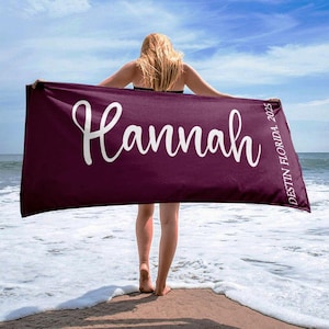 Puede incluir: Una toalla de playa granate con el nombre "Hannah" en escritura blanca. El texto "Destin Florida 2025" est&aacute; impreso verticalmente en el lado derecho. La toalla se sostiene en una playa con el oc&eacute;ano al fondo.
