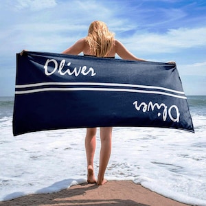 Może przedstawiać: Granatowy ręcznik plażowy z napisem "Oliver" w białym skrypcie. Ręcznik ma dwa poziome białe paski. Osoba trzyma ręcznik na piaszczystej plaży z morzem w tle.