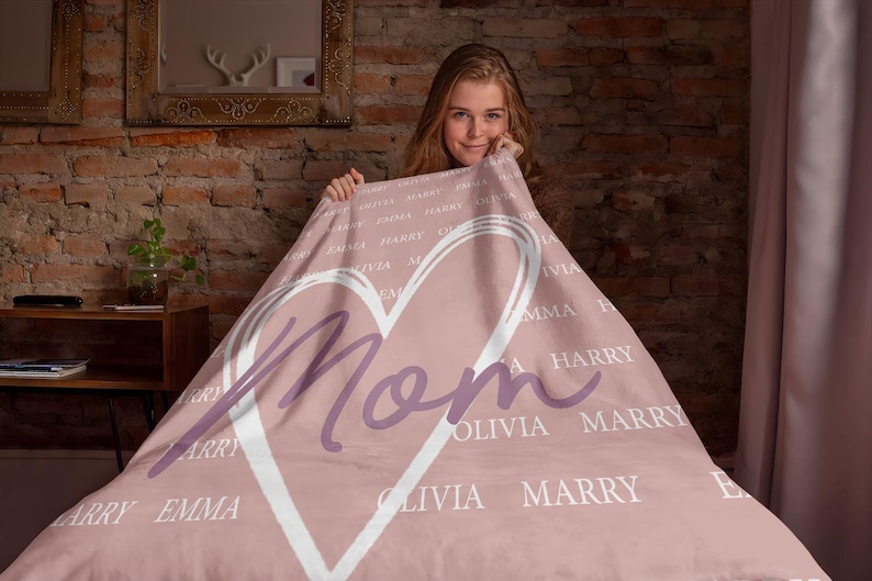 Może przedstawiać: R&oacute;żowy koc polarowy z białym sercem i napisem "Mama" w fioletowym kolorze. Serce jest otoczone imionami w białym kolorze.