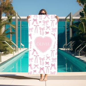 Puede incluir: Toalla de playa blanca con un dise&ntilde;o de coraz&oacute;n rosa y el nombre "Olivia" en el centro. La toalla est&aacute; cubierta de lazos rosas.