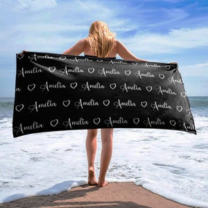 May include: Black beach towel with the name "Amelia" repeated in white cursive lettering with a heart between each name.