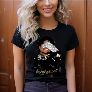 May include: A black short-sleeved t-shirt featuring a graphic print of Michael Jackson. The design shows a portrait of the artist with a sparkling silver sequined glove covering part of the face and the name Michael written in gold cursive script.