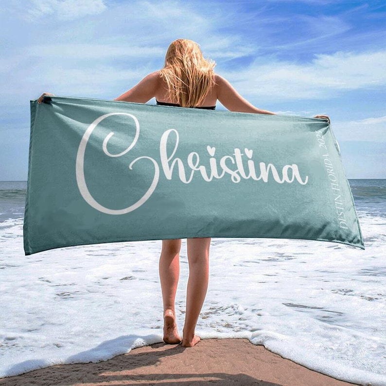 Puede incluir: Una toalla de playa turquesa con el nombre "Christina" en escritura blanca. La toalla es sostenida por una persona en una playa de arena con el oc&eacute;ano al fondo. El texto "Destin Florida 2023" tambi&eacute;n est&aacute; en la toalla.