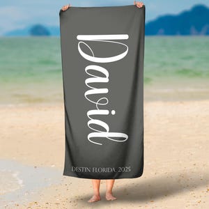 Puede incluir: Una toalla de playa gris oscuro con el nombre "David" en letras grandes, blancas y cursivas. El texto "DESTIN FLORIDA 2025" est&aacute; impreso en la parte inferior. La toalla se muestra sobre un fondo de playa y oc&eacute;ano.