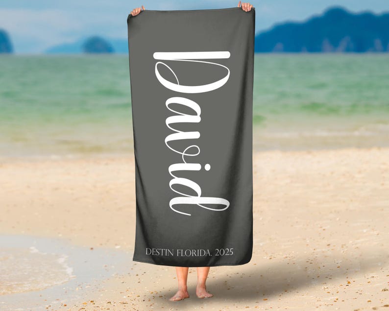 Puede incluir: Una toalla de playa gris oscuro con el nombre "David" en letras grandes, blancas y cursivas. El texto "DESTIN FLORIDA 2025" est&aacute; impreso en la parte inferior. La toalla se muestra sobre un fondo de playa y oc&eacute;ano.