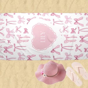 Puede incluir: Toalla de playa rosa con fondo blanco y un coraz&oacute;n rosa con el nombre "LILY" en blanco. La toalla est&aacute; cubierta de un patr&oacute;n de lazos rosas.