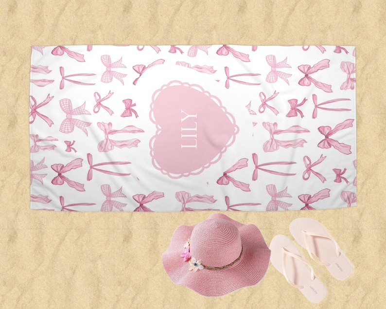 Puede incluir: Toalla de playa rosa con fondo blanco y un coraz&oacute;n rosa con el nombre "LILY" en blanco. La toalla est&aacute; cubierta de un patr&oacute;n de lazos rosas.