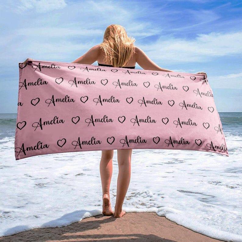 May include: A pink beach towel with the name "Amelia" repeated in black cursive lettering with hearts between each name.
