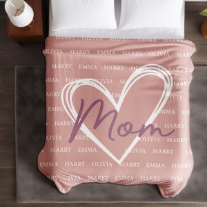 Może przedstawiać: R&oacute;żowy koc z białym sercem i napisem "Mom" w fioletowym kolorze. Koc jest pokryty powtarzającym się wzorem z imionami, w tym "Harry", "Olivia", "Marry" i "Emma".