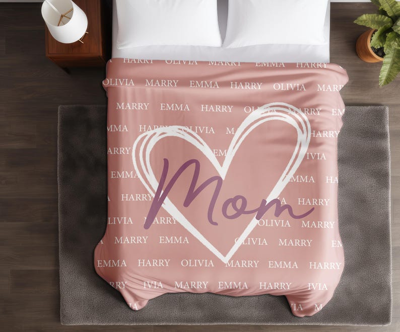Może przedstawiać: R&oacute;żowy koc z białym sercem i napisem "Mom" w fioletowym kolorze. Koc jest pokryty powtarzającym się wzorem z imionami, w tym "Harry", "Olivia", "Marry" i "Emma".